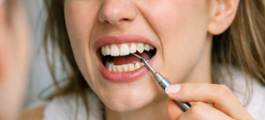 Woman attempting to remove tartar from upper and lower teeth using a metal dental pick, wearing a white bathrobe and headband, with a strained expression in a blurred bathroom setting